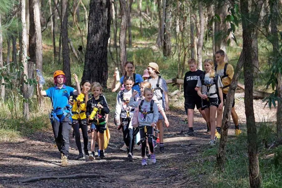 A group of children with an adult guide, wearing outdoor gear and helmets, hiking on a forest trail in daylight.