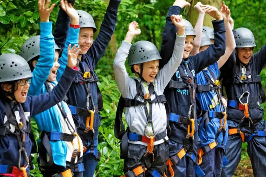 A group of children wearing helmets and harnesses joyfully raising their arms in a forest setting.