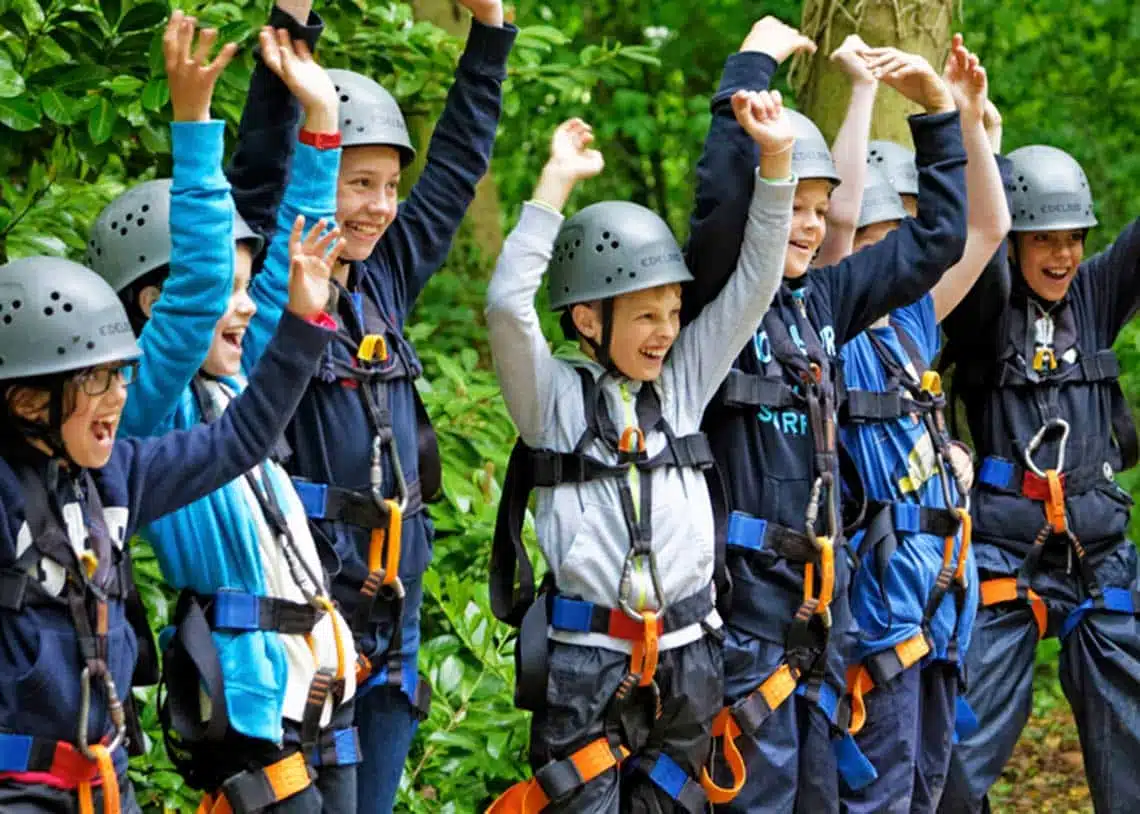 A group of children wearing helmets and harnesses joyfully raising their arms in a forest setting.