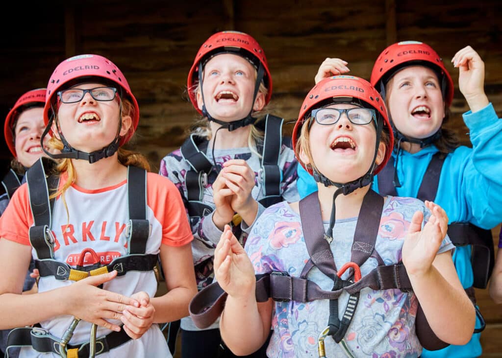 Group of young girls wearing helmets and harnesses, laughing and looking upwards with excitement.