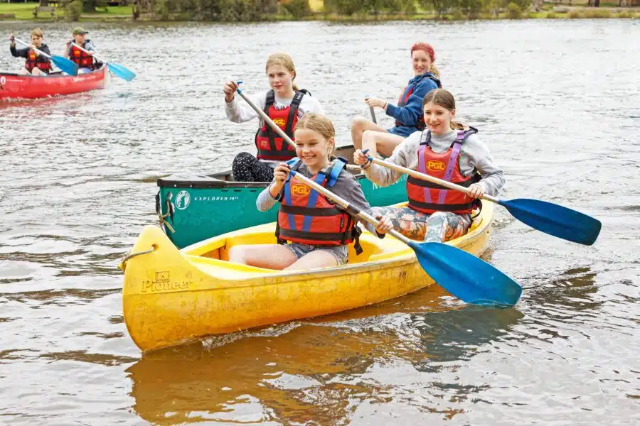 Group of smiling children wearing life jackets paddling a yellow canoe on a river, with other kids canoeing in the background.