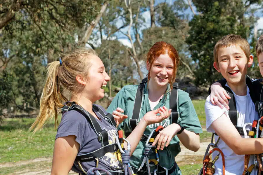 Four children with climbing gear smile and huddle together in a sunny, tree-filled park.