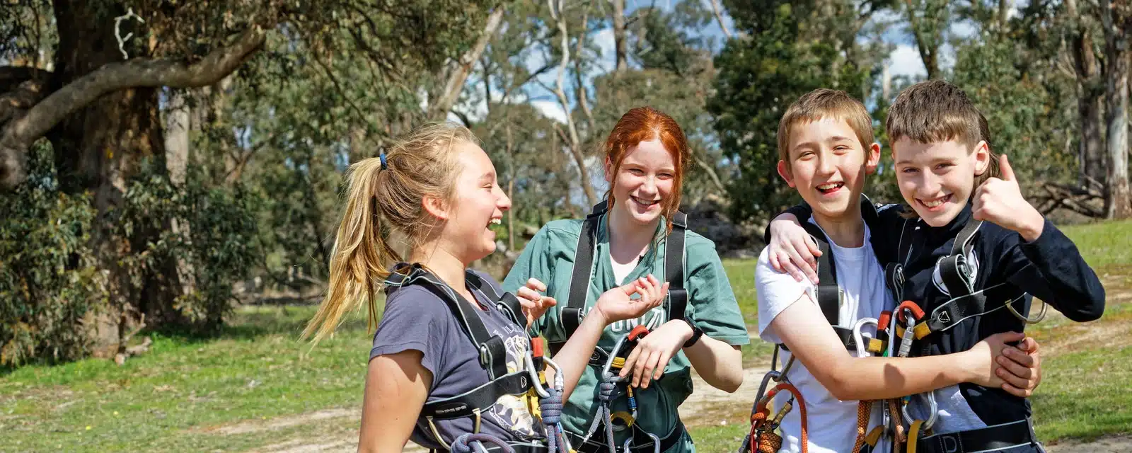 Four children with climbing gear smile and huddle together in a sunny, tree-filled park.