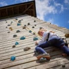 Young girl in a harness climbs a wooden climbing wall under a clear blue sky.