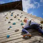 Young girl in a harness climbs a wooden climbing wall under a clear blue sky.