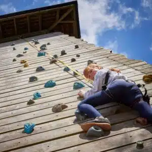 Young girl in a harness climbs a wooden climbing wall under a clear blue sky.