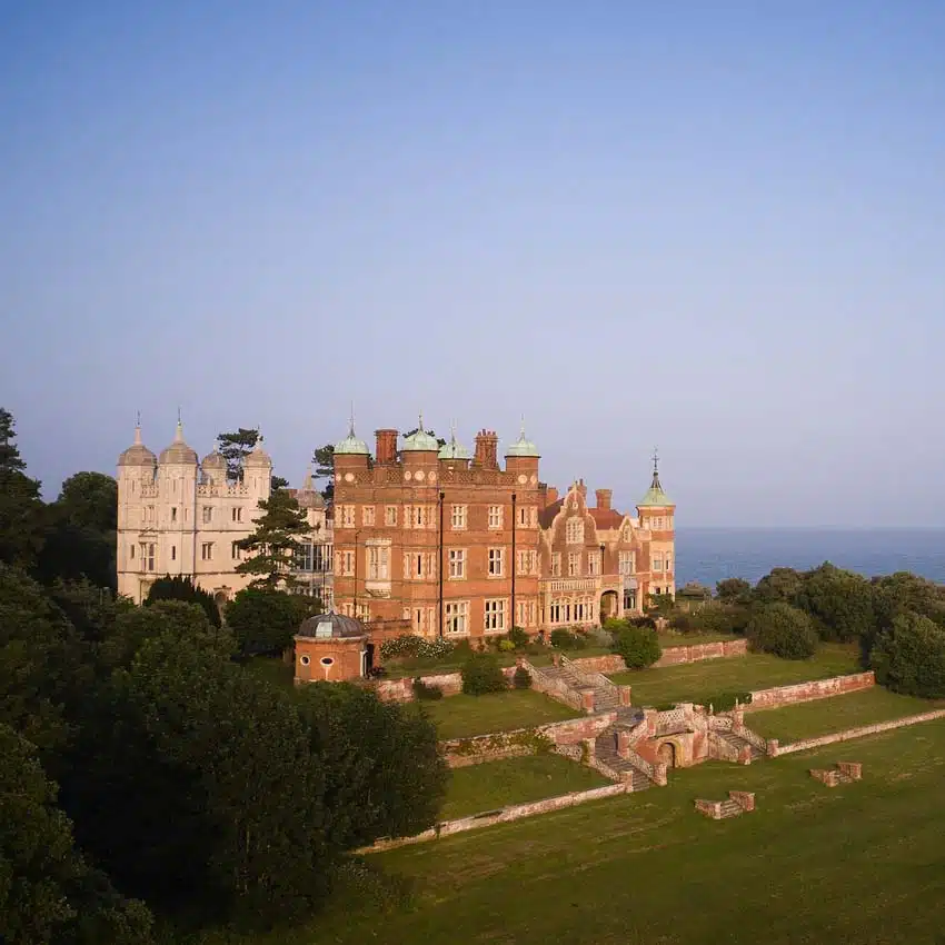 Aerial view of a grand historic mansion with ornate architecture, surrounded by green lawns and lush trees at sunset.