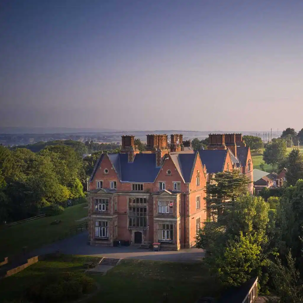 Aerial view of an expansive, red-brick Victorian mansion surrounded by lush green lawns and clear skies during early evening.