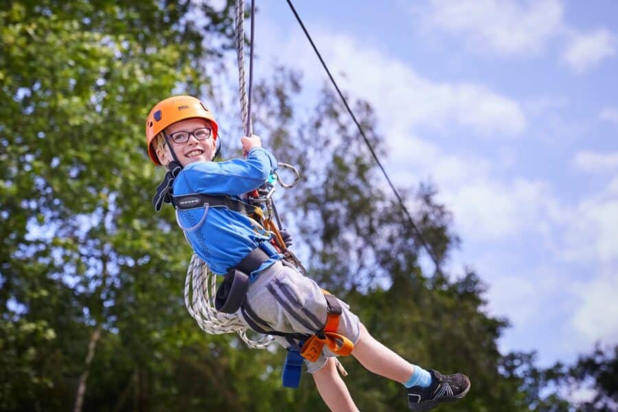 Young boy with glasses and helmet joyfully zip-lining against a backdrop of trees and a clear sky, enjoying the physical activity benefits for kids.