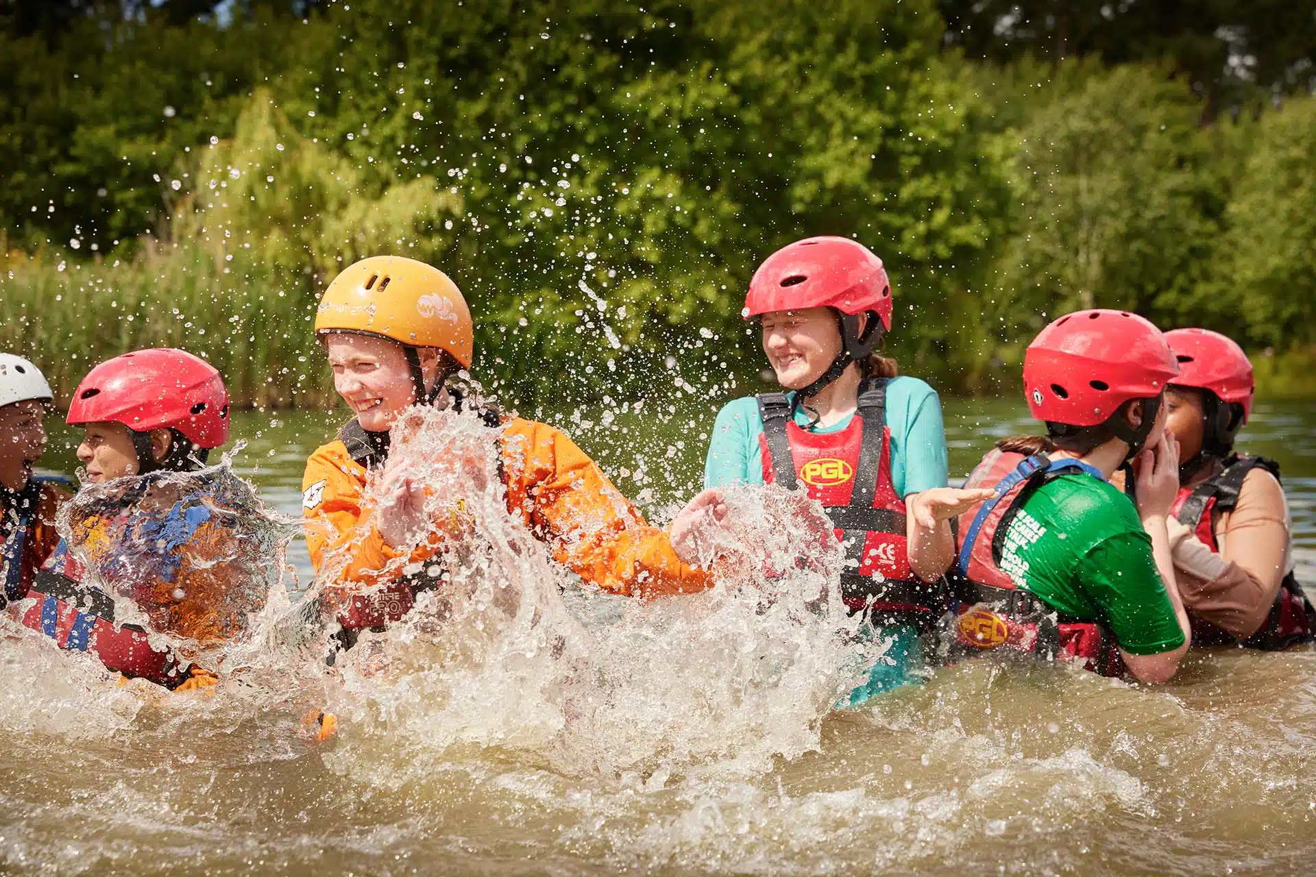 Group of young people wearing helmets and life jackets laughing and splashing water while rafting on a river.