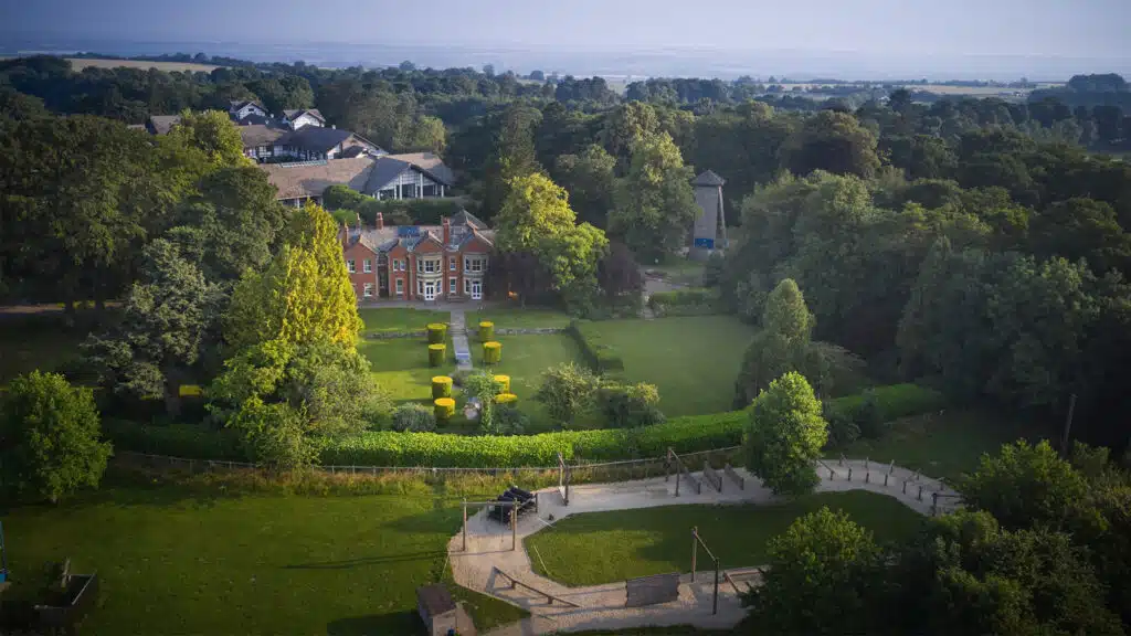 Aerial view of Newby Wiske Hall, featuring a large estate with manicured gardens and a brick house, surrounded by lush greenery and distant rolling hills.