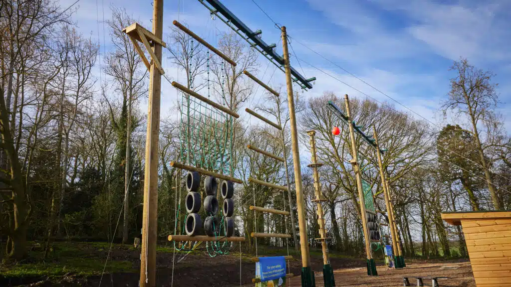 Outdoor adventure playground at Newby Wiske Hall with various climbing obstacles including ropes, wooden beams, and tire swings, set against a clear blue sky and surrounded by trees.