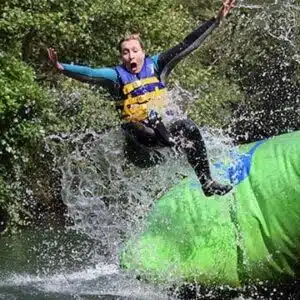 A person in a wetsuit and life jacket leaps off a green inflatable slide at Newby Wiske Hall into a splash of water.