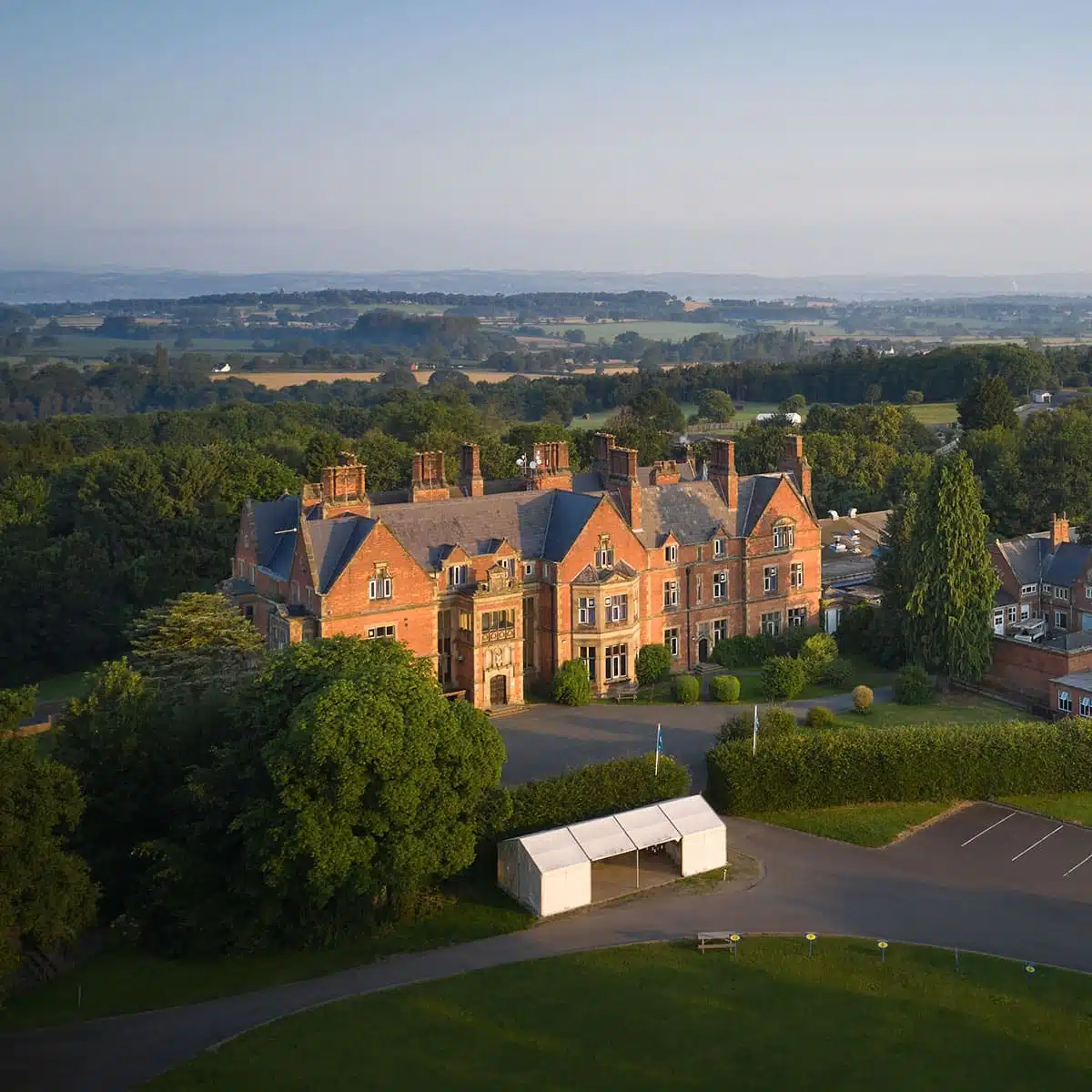 Aerial view of an ornate brick mansion surrounded by lush green lawns and trees, with a hazy countryside backdrop at sunrise.