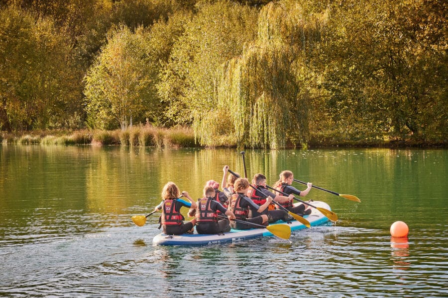 Group of people in life jackets paddling a large raft on a calm lake with lush green trees in the background.