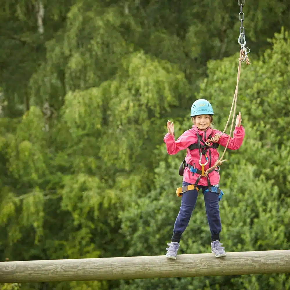 A young child in a helmet and harness smiles while balancing on a log during a zip line adventure in a forested area.