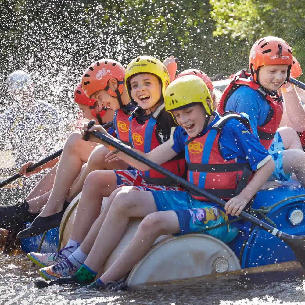 Group of young people wearing helmets and life jackets, enjoying a splash-filled raft ride on a sunny day.
