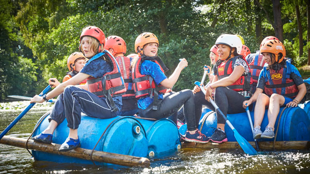 Group of children in helmets and life jackets laughing and paddling together on a raft made of blue barrels on a river.