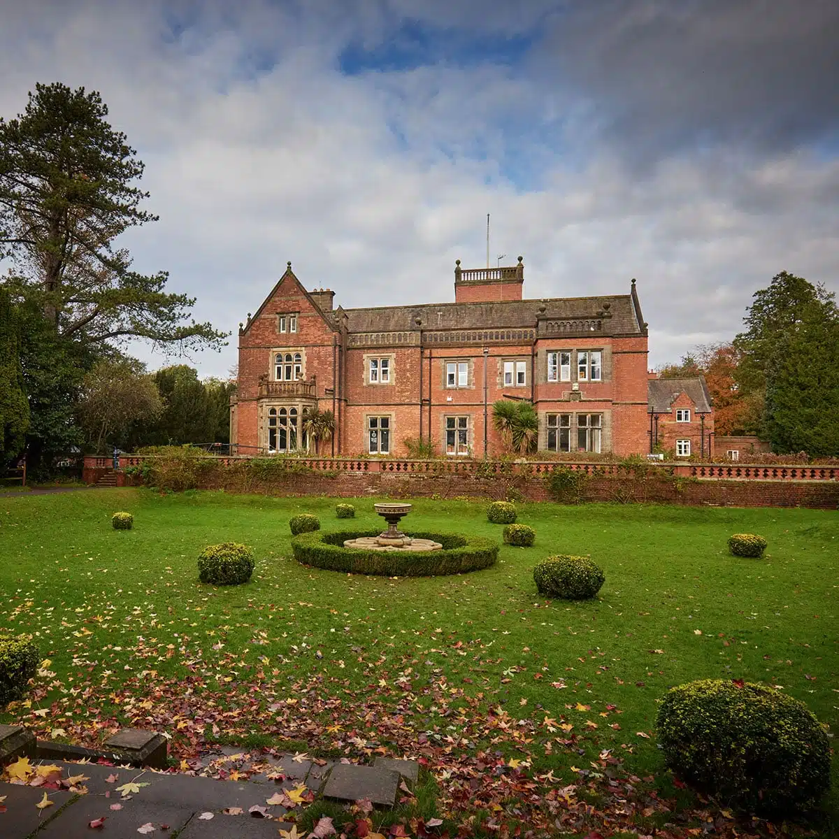 Historic red brick mansion with ornate windows, surrounded by a lush garden and a central fountain, under a cloudy sky.