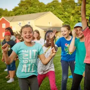 Children celebrating outdoors, with one girl in a blue shirt leading and others cheering and smiling in the background.