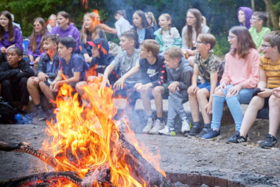 A group of young campers sits around a large campfire in a forest clearing, watching the flames intently.