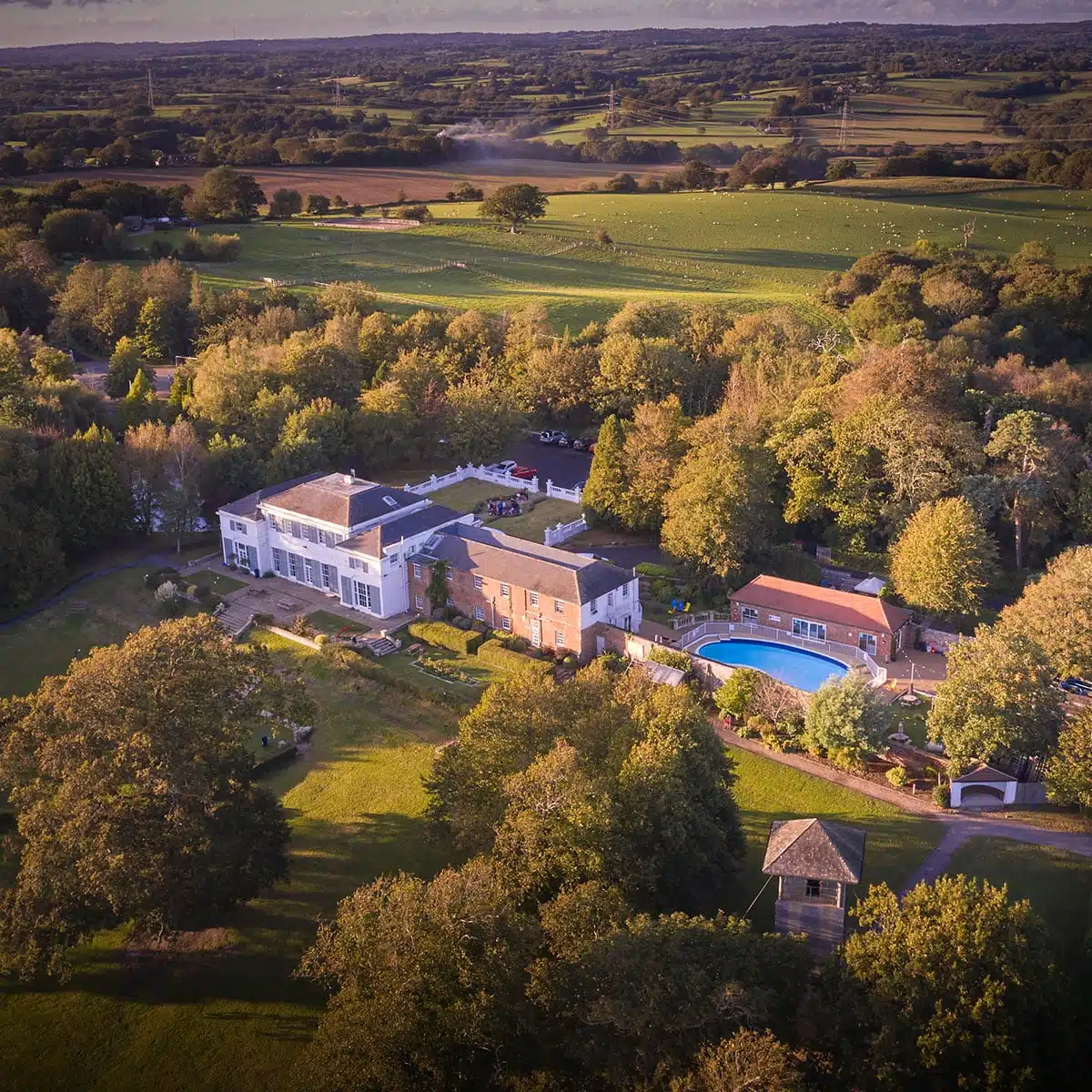 Aerial view of a luxurious estate with multiple buildings and a swimming pool, surrounded by lush greenery and fields at sunset.