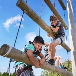 Two children wearing harnesses climb on a wooden outdoor adventure playground under a clear blue sky.