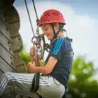 Young boy in a helmet and harness smiles while climbing a rope course outdoors.