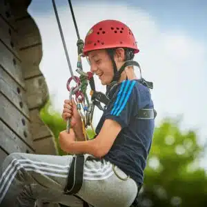 Young boy in a helmet and harness smiles while climbing a rope course outdoors.