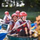 Two young people wearing helmets and life vests paddling a canoe with others in the background, surrounded by green foliage.