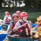 Two young people wearing helmets and life vests paddling a canoe with others in the background, surrounded by green foliage.