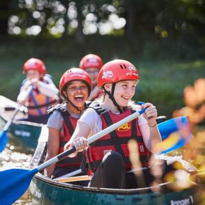 Two young people wearing helmets and life vests paddling a canoe with others in the background, surrounded by green foliage.