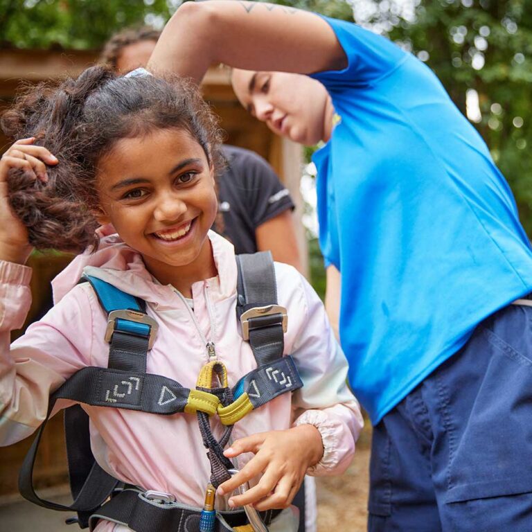 A young girl smiles at the camera while an instructor adjusts her climbing harness in a wooded area.
