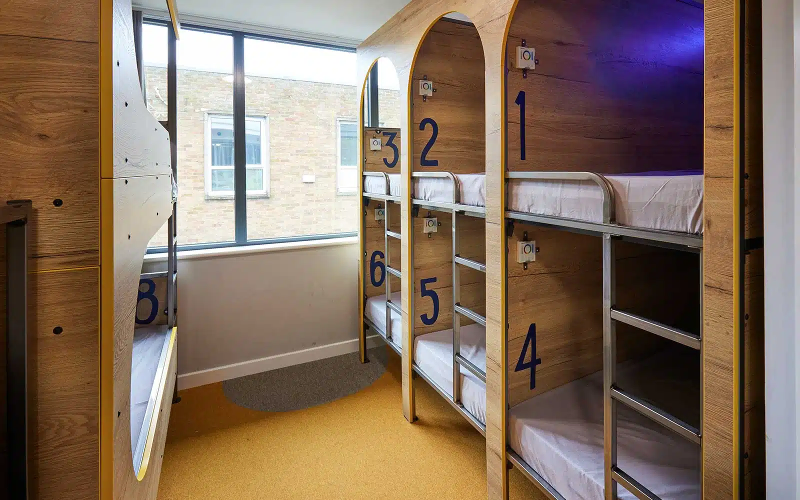 Modern hostel room in Newby Wiske Hall with numbered wooden bunk beds arranged in a circular pattern around a central pillar, with a window on the side.