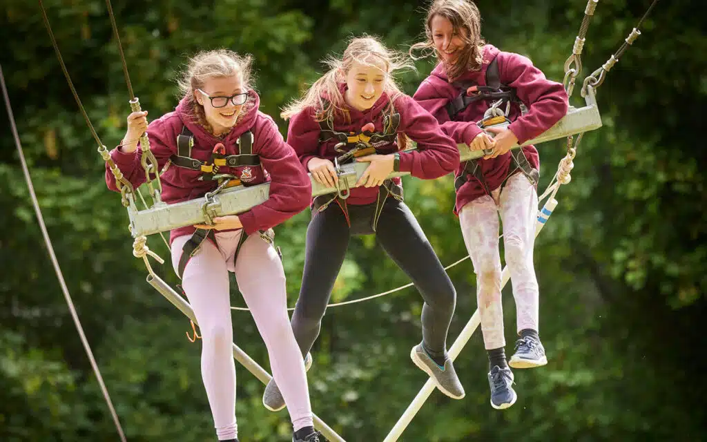Three girls in safety harnesses enjoy a multi-activity rope swing ride outdoors, smiling and holding onto the ropes.