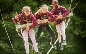 Three girls in safety gear and harnesses joyfully navigate a multi-activity rope course amidst lush greenery.