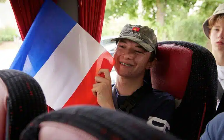 A smiling young person holding the French flag while sitting on a multi-activity bus, wearing a cap and a backpack.