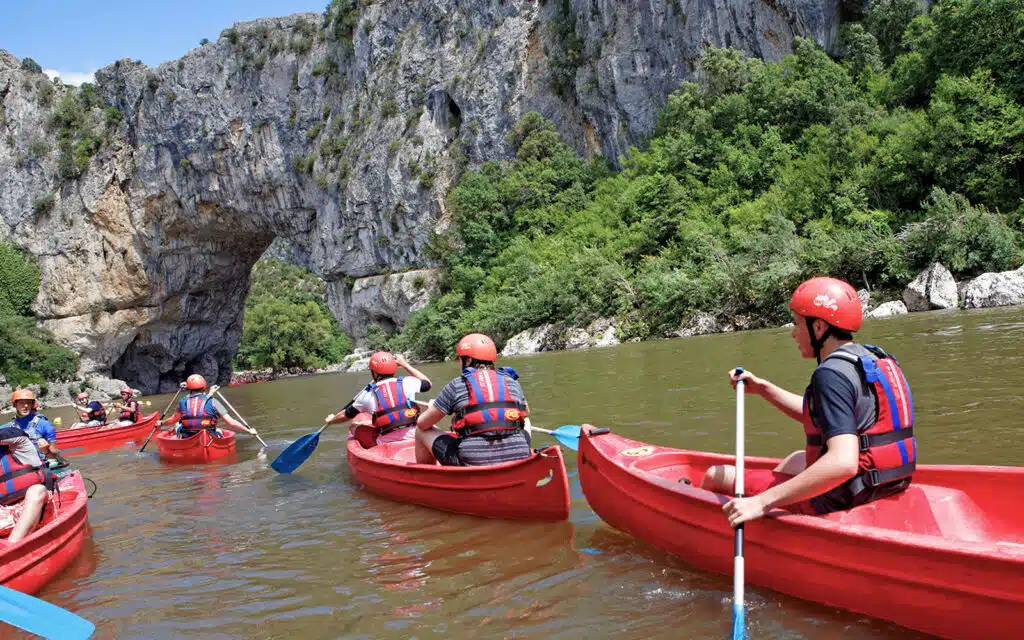 People in red kayaks wearing helmets and life jackets engage in multi-activity paddle sports on a river near a large rock arch formation.