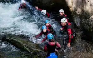 A group of six people with helmets and life jackets engage in a multi-activity canyoning adventure, navigating a rocky, fast-flowing stream.