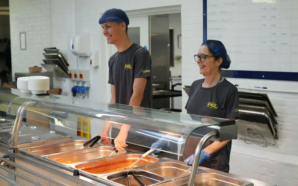 Two cafeteria workers serving food, smiling in a clean, organized kitchen with trays and food warmers.