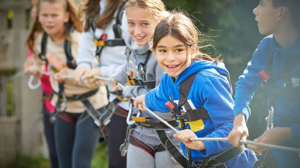 A group of young teens in safety harnesses engaging in an outdoor rope pulling activity at PGL Adventure Holidays, smiling and focused.