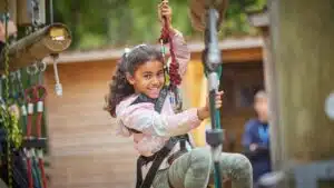 A young girl with a joyful expression glides on a zipline at an outdoor adventure park, surrounded by ropes and harnesses.