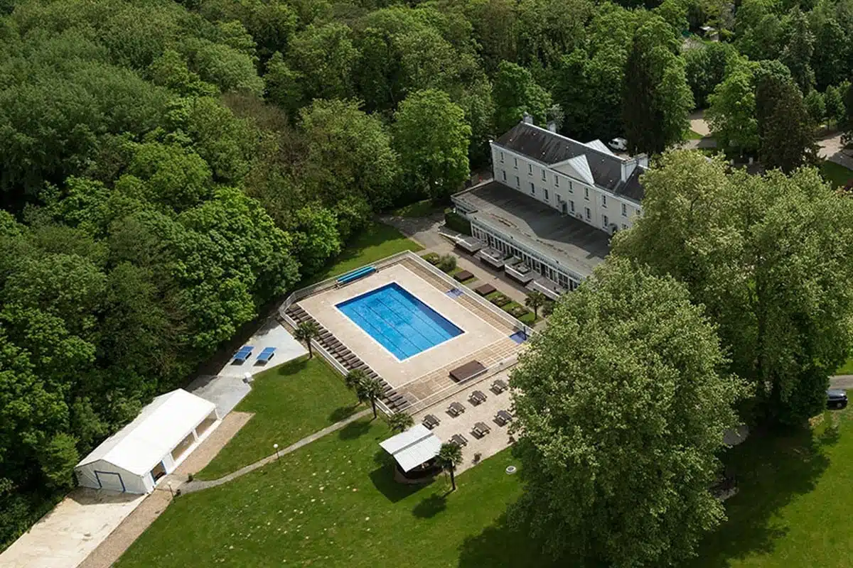 Aerial view of a large estate with a white building and an outdoor swimming pool surrounded by dense green trees.