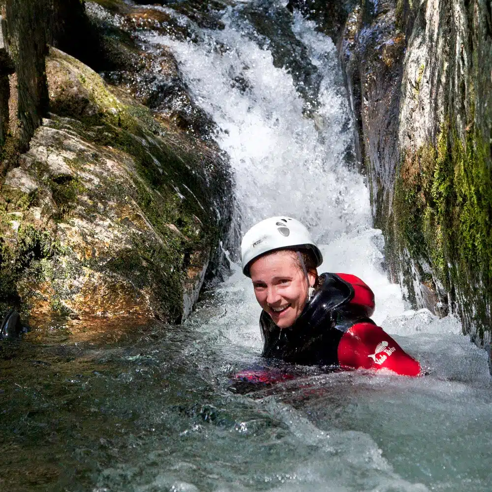 A person wearing a white helmet and red life jacket smiles while swimming down a rocky waterfall.