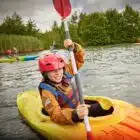 A child in a yellow kayak wears a red helmet and life jacket while paddling on a lake surrounded by greenery.
