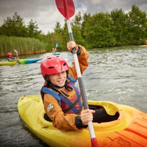 A child in a yellow kayak wears a red helmet and life jacket while paddling on a lake surrounded by greenery.