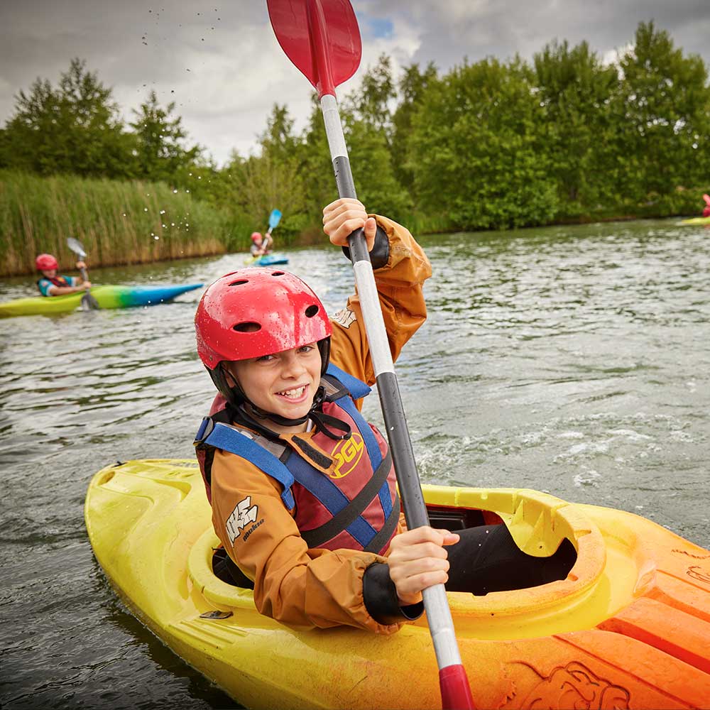 A child in a yellow kayak wears a red helmet and life jacket while paddling on a lake surrounded by greenery.