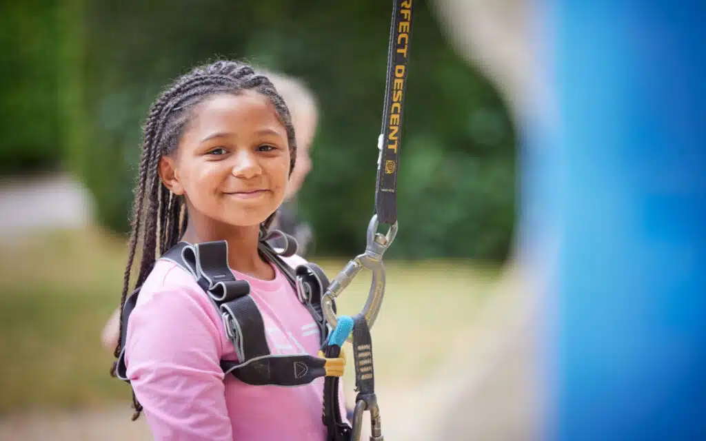 A young girl with braided hair, wearing a harness, smiles at the camera, ready for a climbing activity.