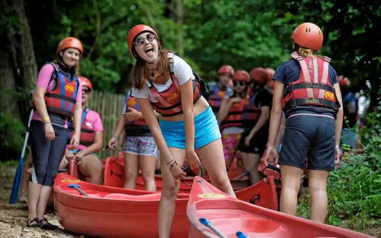 A group of people in helmets and life jackets laughing near red kayaks, getting ready for a multi-activity river trip.