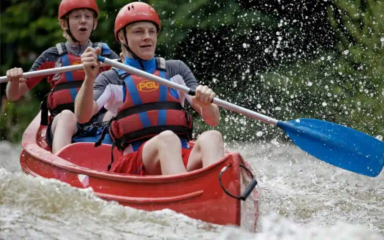 Two people in red life vests engaging in a multi-activity adventure, paddling a canoe through splashing water.